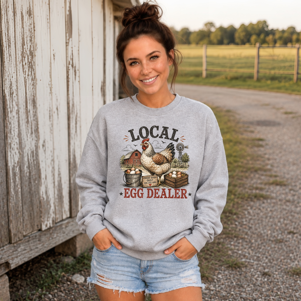 Woman wearing a sports gray Local Egg Dealer sweatshirt outside a weathered barn.