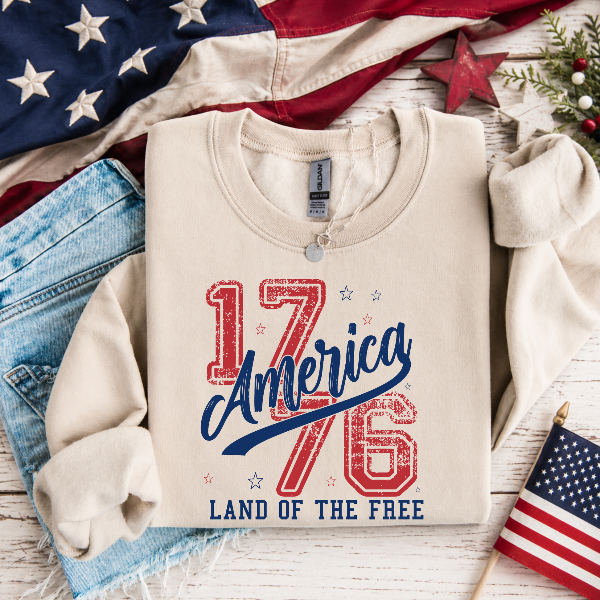Beige sweatshirt with '1776 America Land of the Free' text, American flag, jeans, and small flags on a wooden surface.