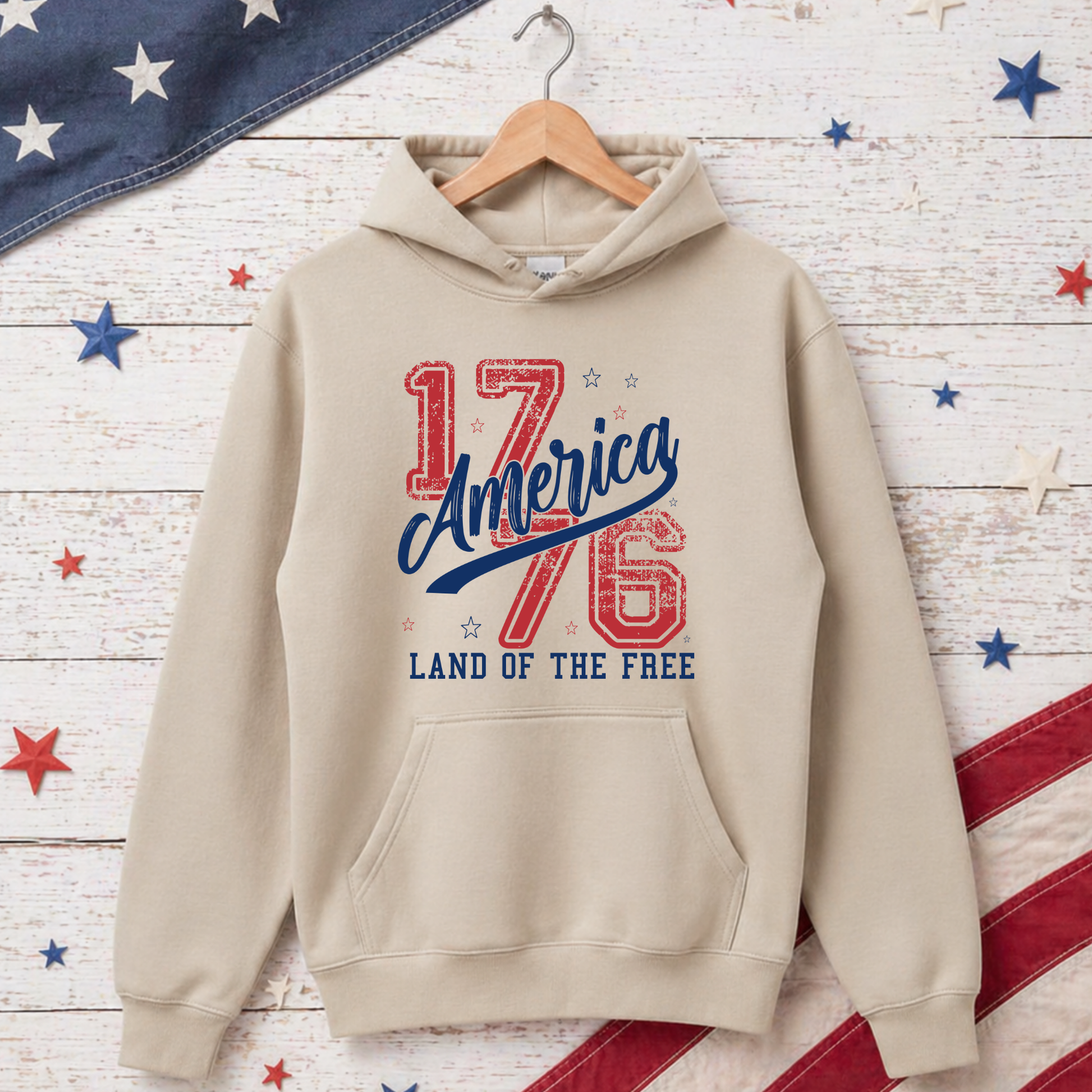 Beige hoodie with 'America 1776 Land of the Free' text on a wooden background with American flag decorations.