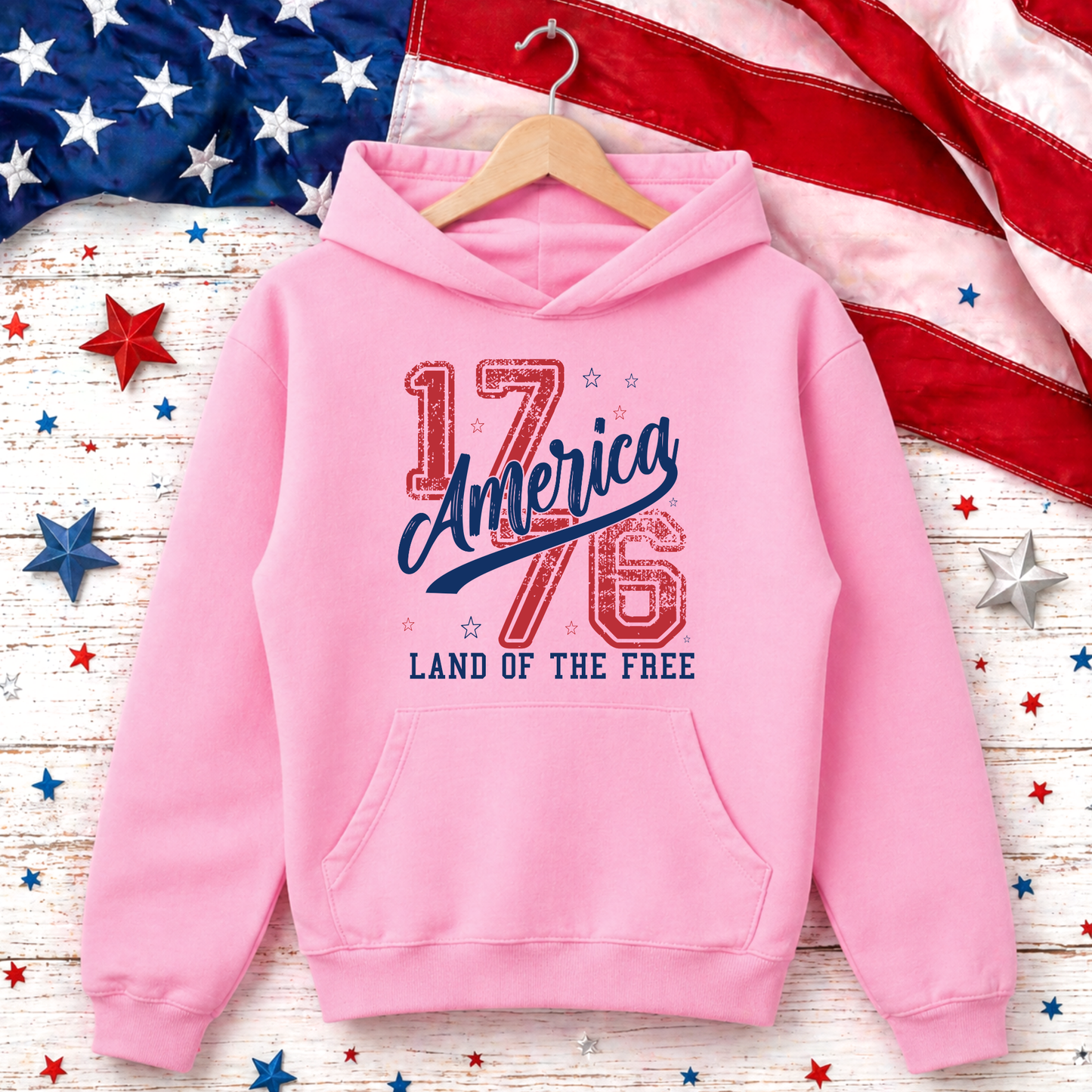 Pink hoodie with 'America 1776 Land of the Free' text on a wooden surface with American flag and stars.