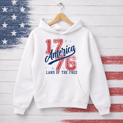 White hoodie with '1776 America Land of the Free' text on a wooden background with American flag pattern
