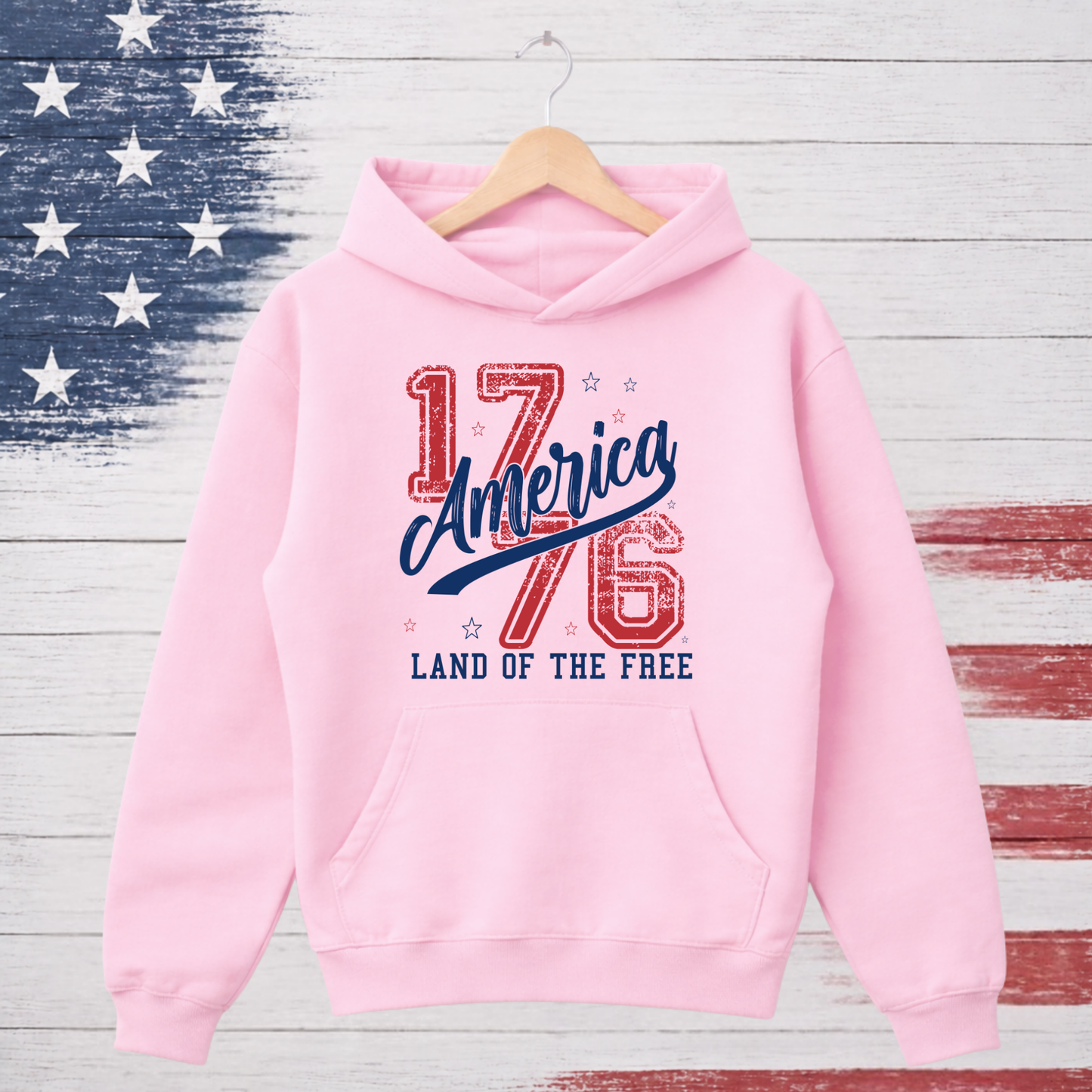 Pink hoodie with 'America 1776 Land of the Free' text on a wooden background with American flag design.