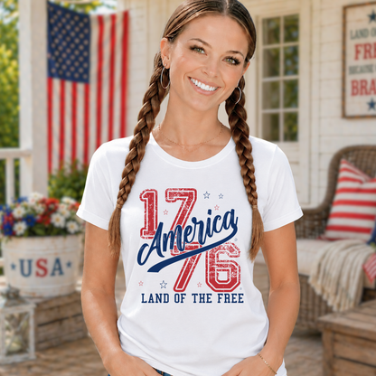 Woman wearing a white t-shirt with '1776 America Land of the Free' design on a porch with an American flag in the background.