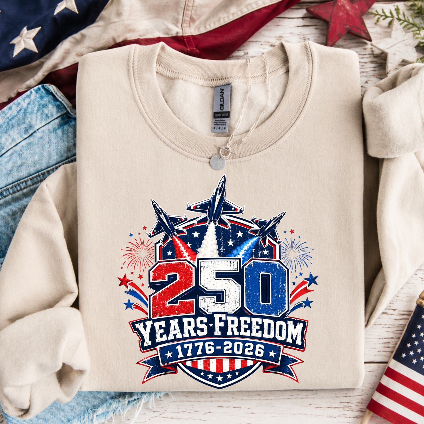 Beige sweatshirt with '250 Years Freedom 1776-2026' design on a wooden surface with American flags and jeans.