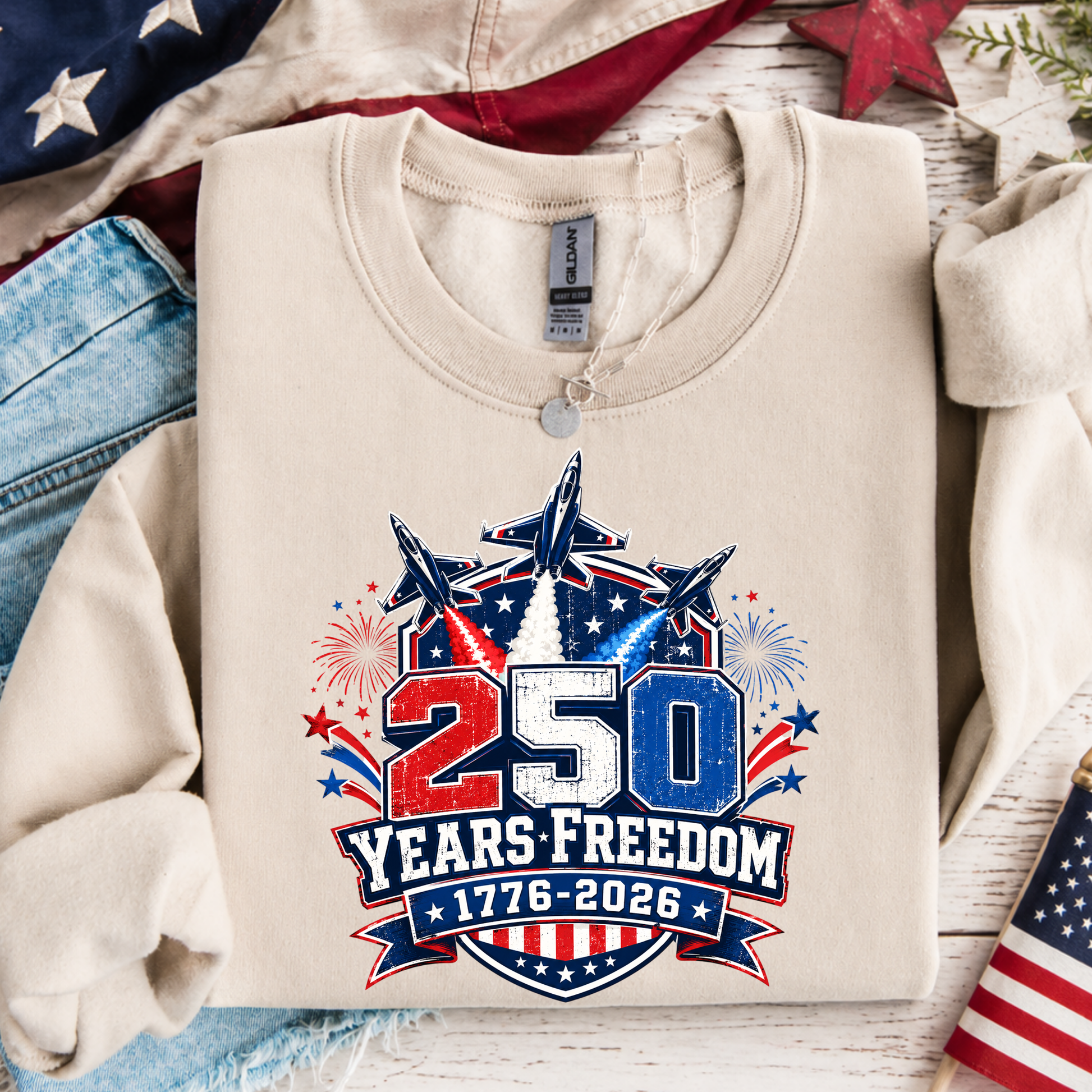 Beige sweatshirt with '250 Years Freedom 1776-2026' design on a wooden surface with American flags and jeans.