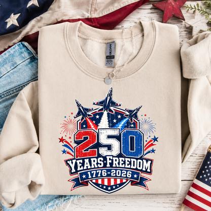Beige sweatshirt with '250 Years Freedom 1776-2026' design on a wooden surface with American flags and jeans.