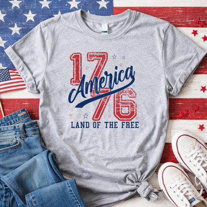 Gray t-shirt with '1776 America Land of the Free' design on a wooden surface with American flags and shoes.