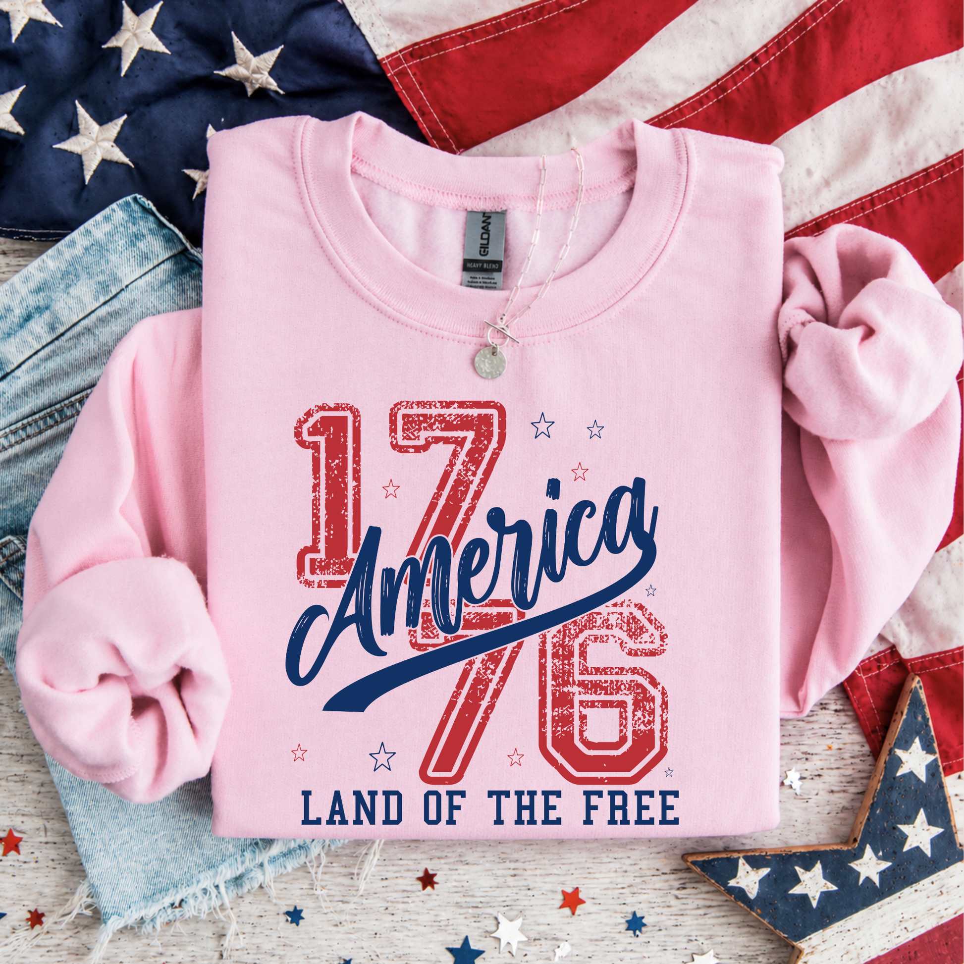 Pink shirt with 'America 1776 Land of the Free' text on a wooden surface with American flags.