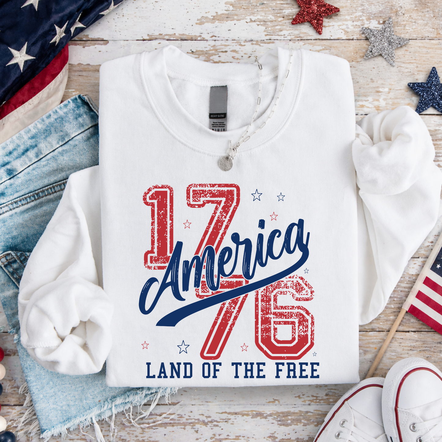 White sweatshirt with '1776 America Land of the Free' text on a wooden surface with American flags and denim jeans.