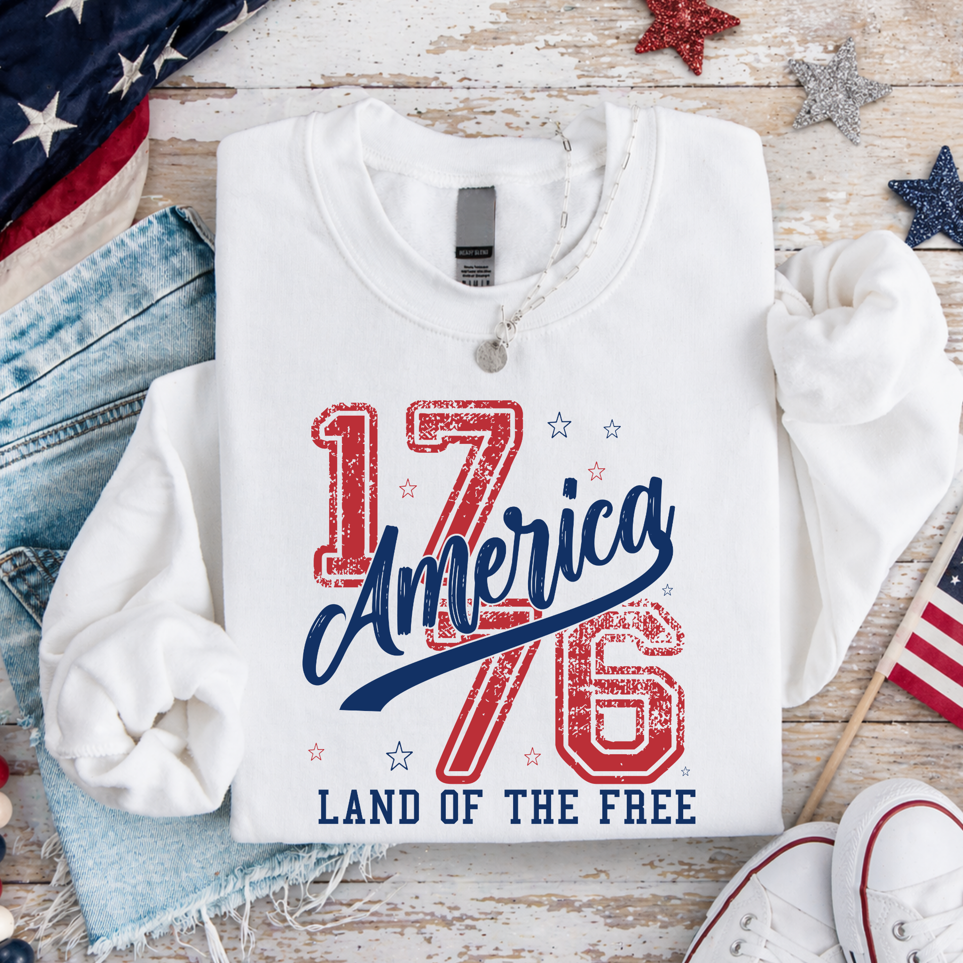 White sweatshirt with '1776 America Land of the Free' text on a wooden surface with American flags and denim jeans.