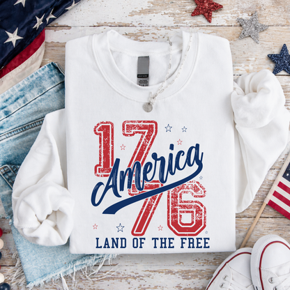 White sweatshirt with '1776 America Land of the Free' text on a wooden surface with American flags and denim jeans.