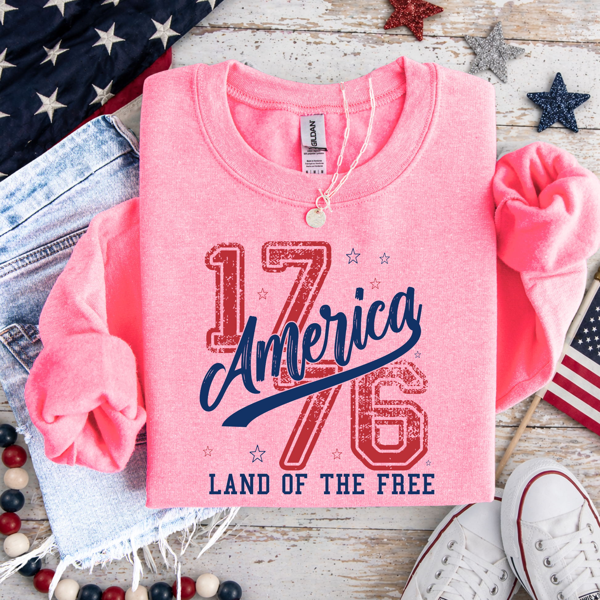 Pink sweatshirt with 'America 1776 Land of the Free' text on a wooden surface with American flags and shoes.