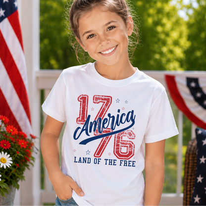 Young girl wearing a white t-shirt with '1776 America Land of the Free' design, standing outdoors with an American flag in the background.