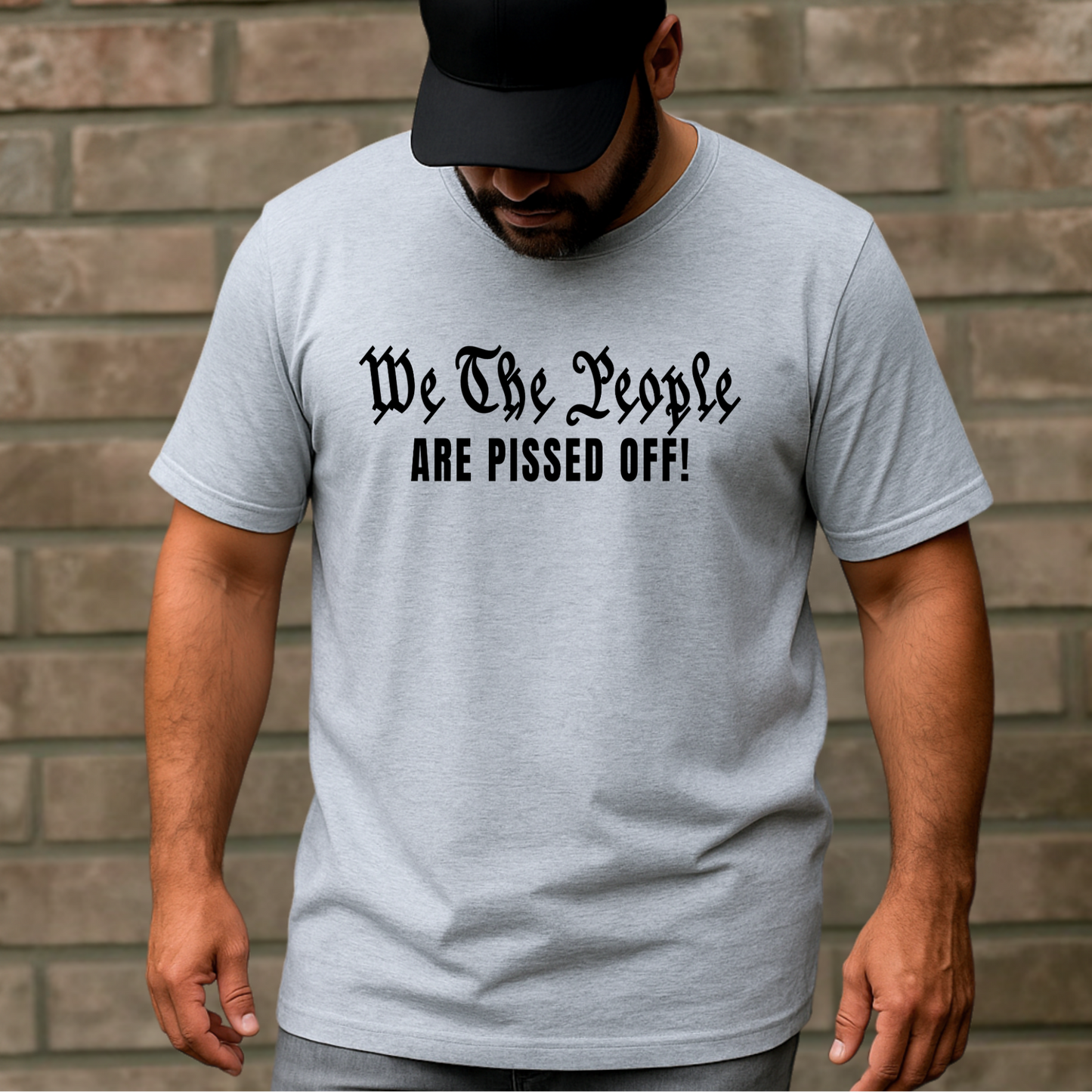 Man wearing a gray t-shirt with text 'We The People ARE PISSED OFF!' against a brick wall.