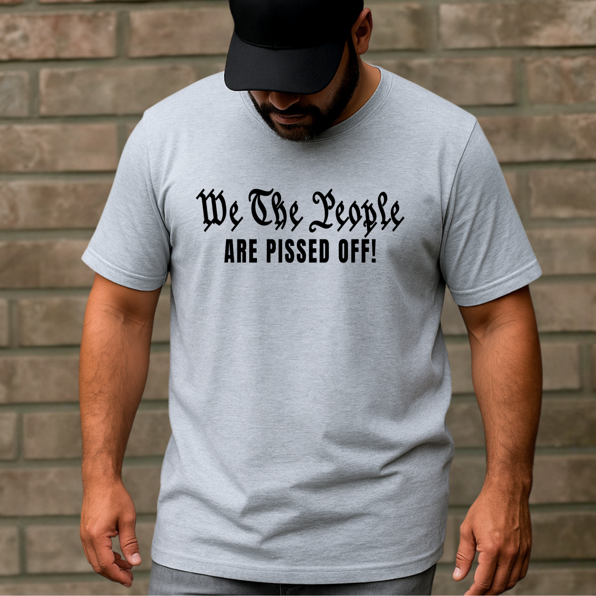 Man wearing a gray t-shirt with text 'We The People ARE PISSED OFF!' against a brick wall.