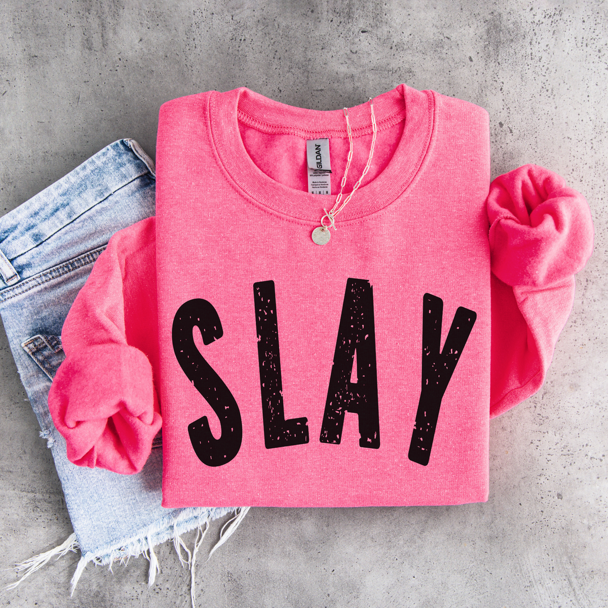 Bright pink SLAY crewneck sweatshirt flat lay with bold black distressed lettering, styled with denim, necklace, and matching scrunchies.