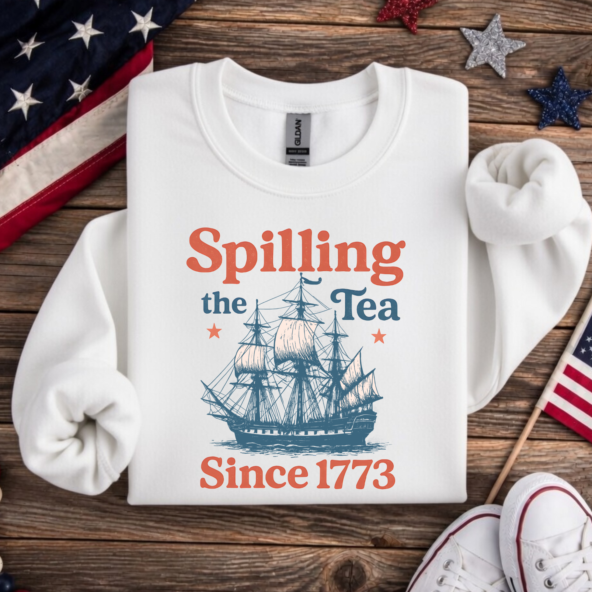 White sweatshirt with ship graphic and text 'Spilling the Tea Since 1773' on a wooden surface with American flags.