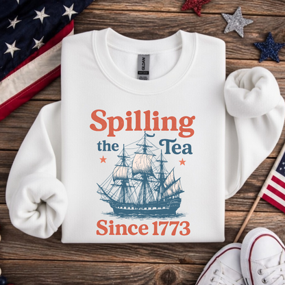 White sweatshirt with ship graphic and text 'Spilling the Tea Since 1773' on a wooden surface with American flags.