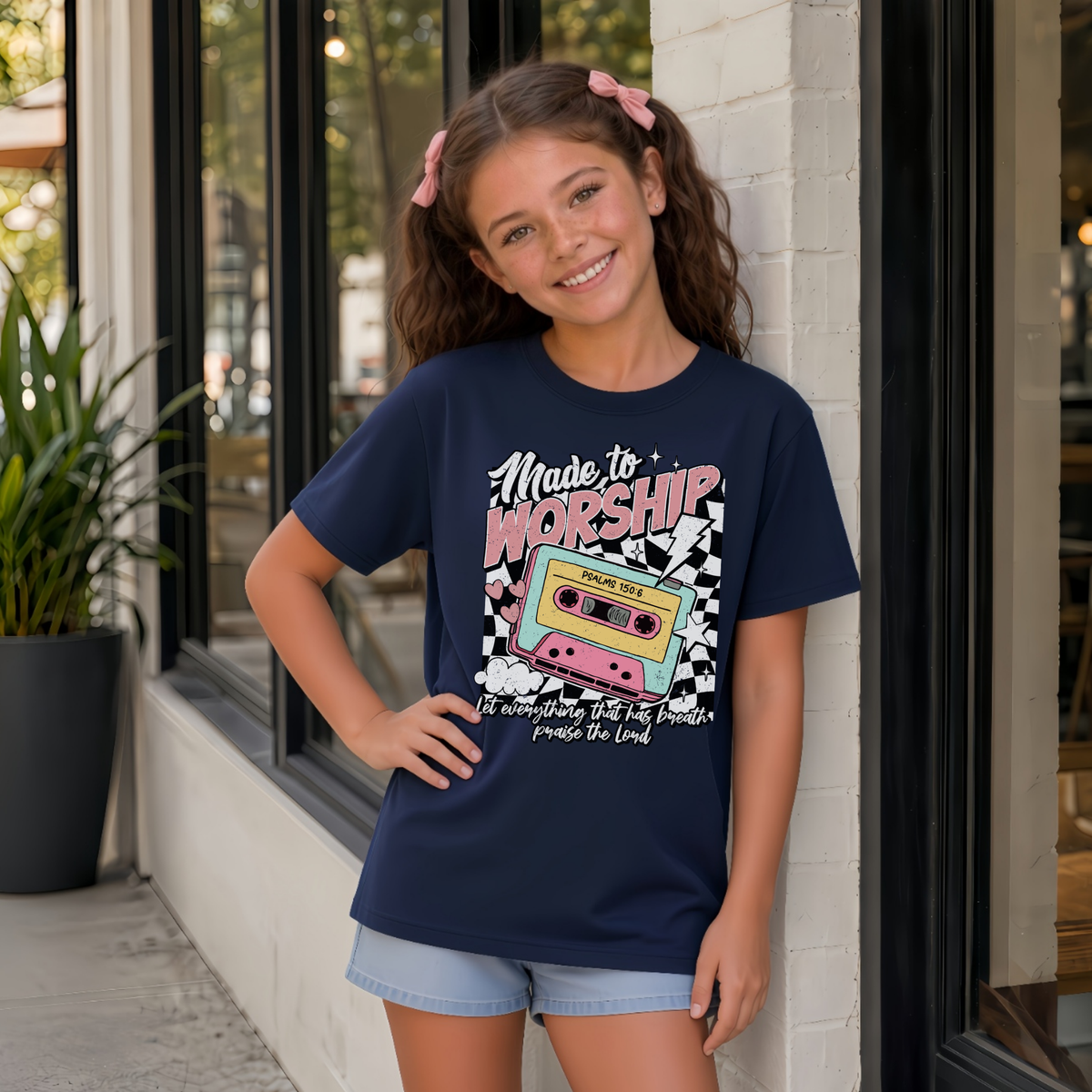Youth girl wearing a navy Made to Worship T-shirt with a colorful retro cassette worship graphic outside a storefront.