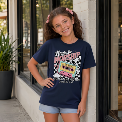 Youth girl wearing a navy Made to Worship T-shirt with a colorful retro cassette worship graphic outside a storefront.