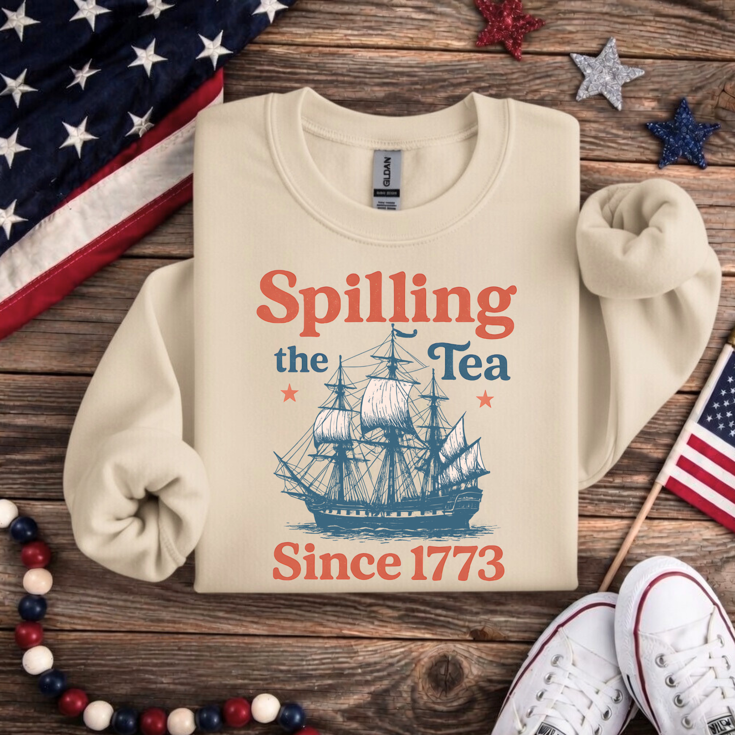 Beige sweatshirt with ship graphic and text 'Spilling the Tea Since 1773' on a wooden surface with American flags.