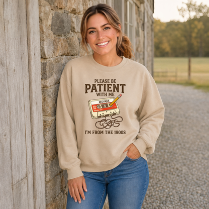 Woman wearing a sand crewneck sweatshirt with the Please Be Patient With Me, I’m From the 1900s cassette design beside a stone wall.