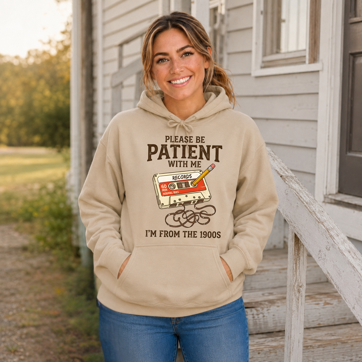 Woman wearing a sand hoodie with a cassette tape and pencil graphic reading Please Be Patient With Me, I’m From the 1900s.