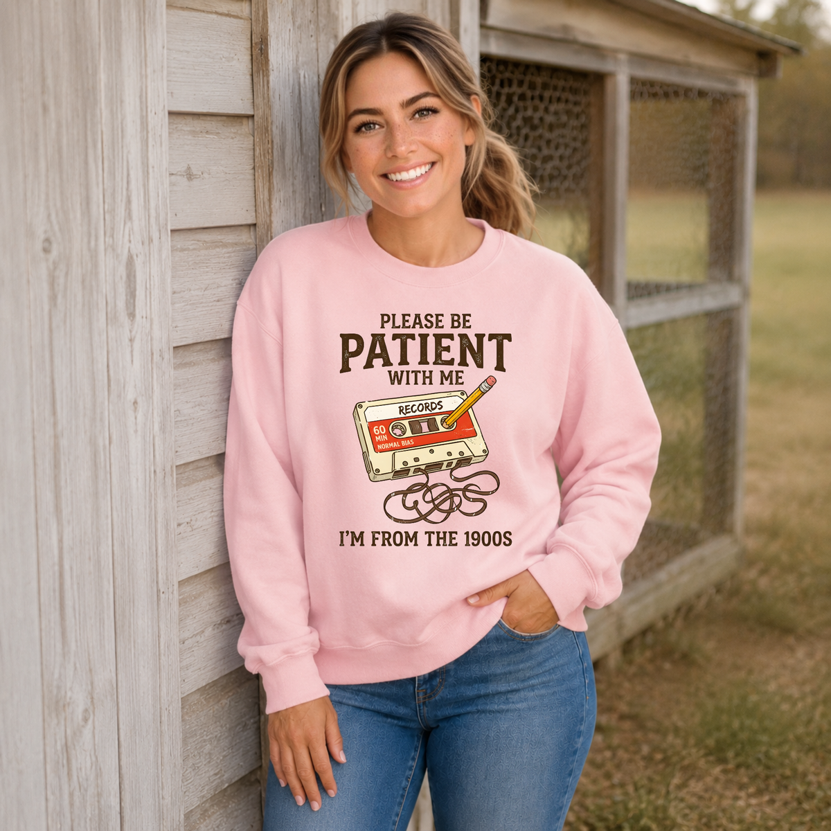 Woman wearing a light pink crewneck sweatshirt featuring the Please Be Patient With Me, I’m From the 1900s cassette design.