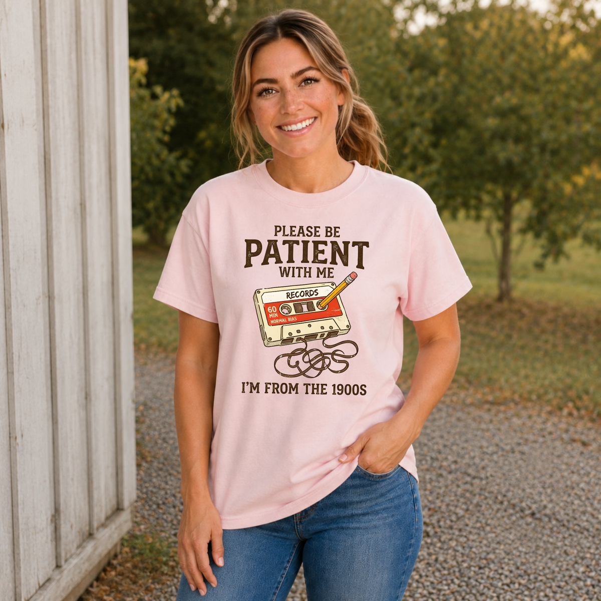 Woman wearing a light pink T-shirt with a cassette tape and pencil design reading Please Be Patient With Me, I’m From the 1900s.