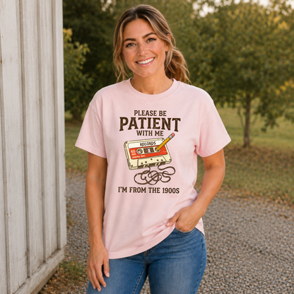 Woman wearing a light pink T-shirt with a cassette tape and pencil design reading Please Be Patient With Me, I’m From the 1900s.