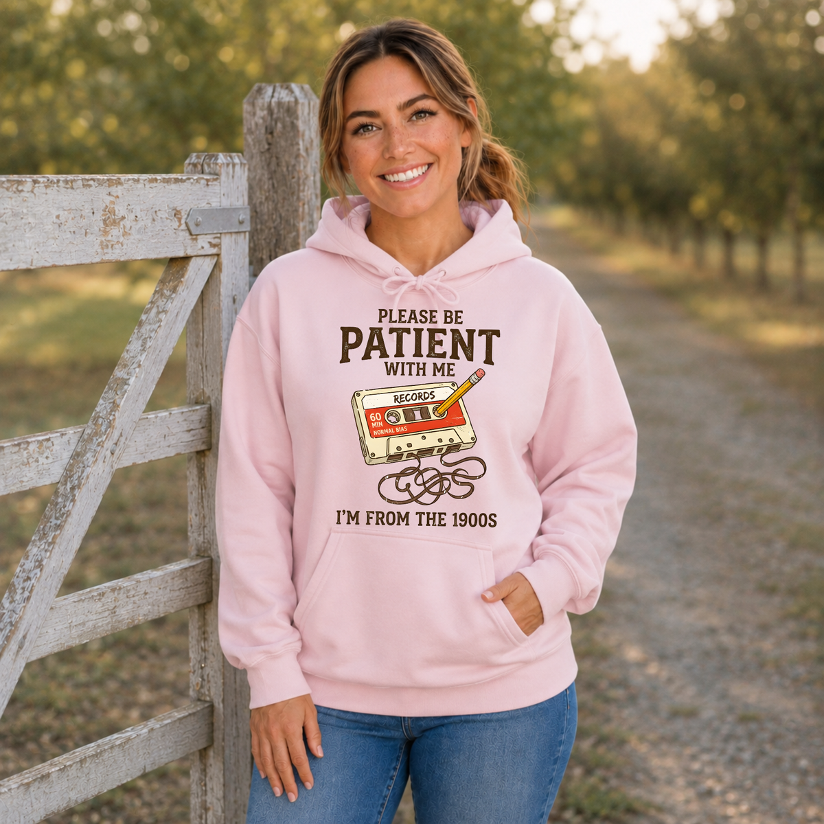 Woman wearing a light pink hoodie featuring the Please Be Patient With Me, I’m From the 1900s cassette tape graphic.