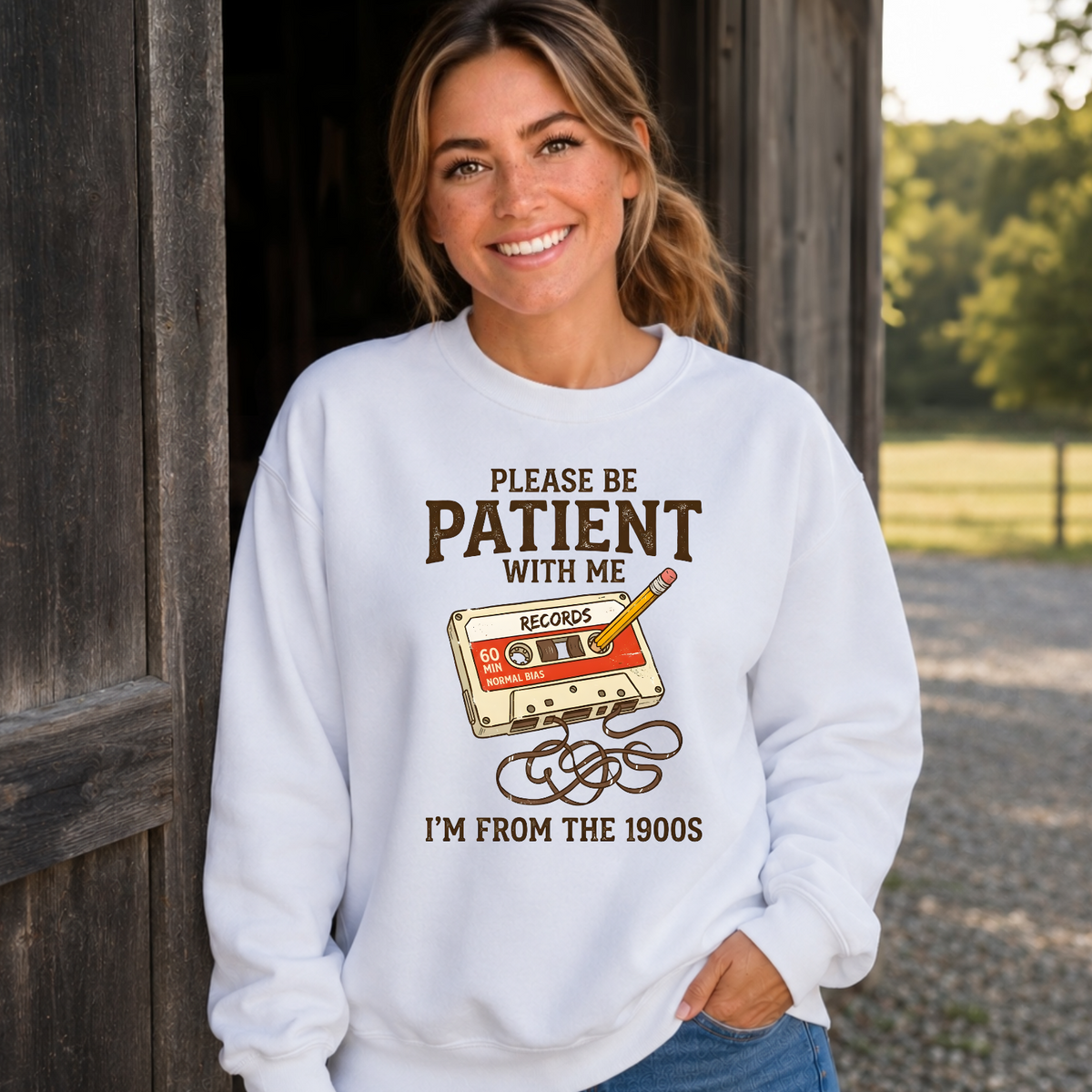 Woman wearing a white crewneck sweatshirt with a cassette tape and pencil graphic reading Please Be Patient With Me, I’m From the 1900s.