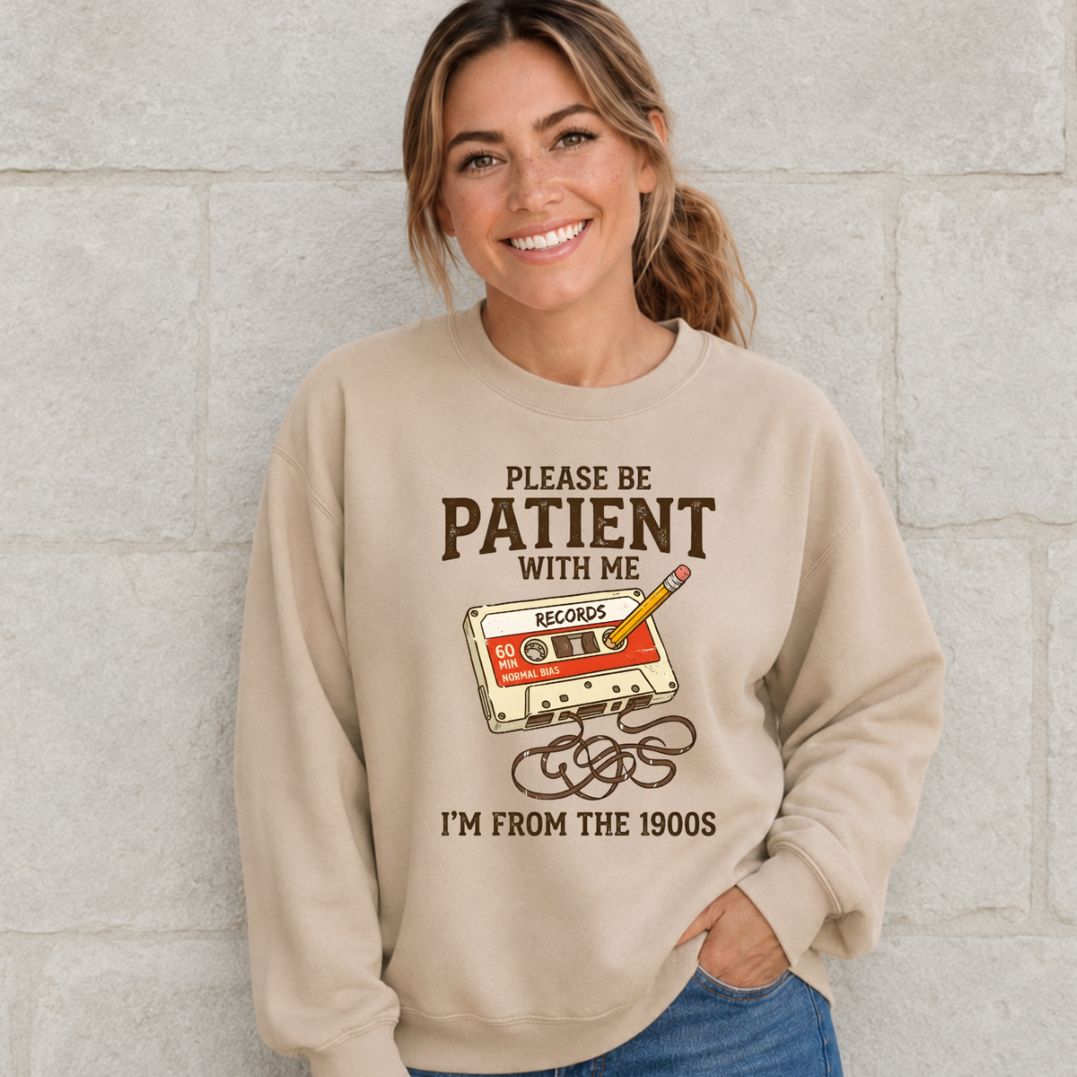 Woman wearing a sand crewneck sweatshirt with a cassette tape and pencil design reading Please Be Patient With Me, I’m From the 1900s.