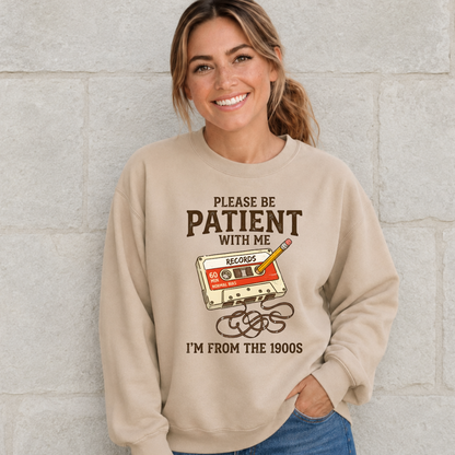 Woman wearing a sand crewneck sweatshirt with a cassette tape and pencil design reading Please Be Patient With Me, I’m From the 1900s.