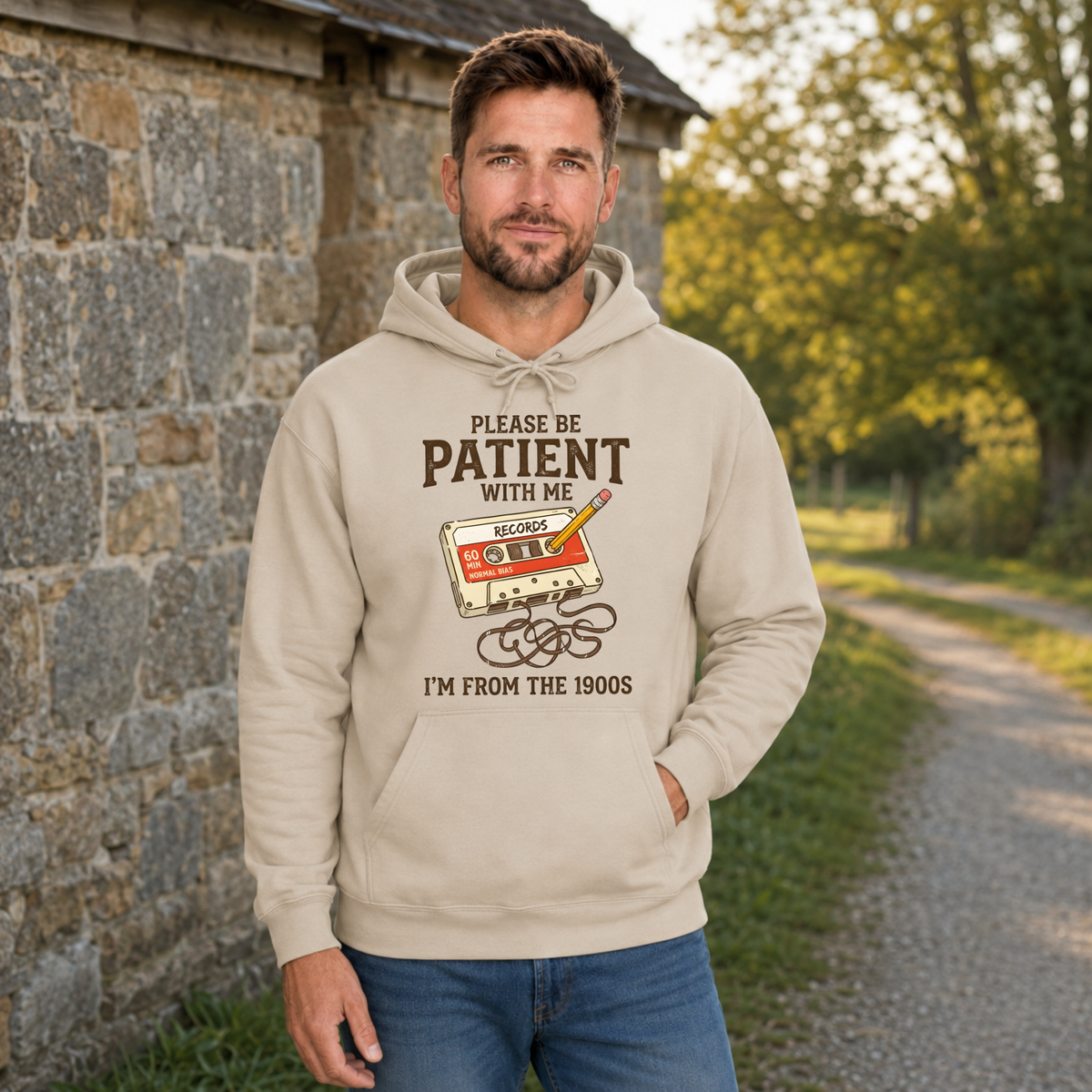 Man wearing a sand hoodie with the Please Be Patient With Me, I’m From the 1900s graphic beside a stone wall.