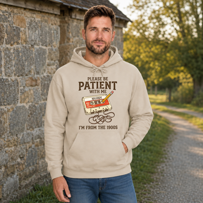 Man wearing a sand hoodie with the Please Be Patient With Me, I’m From the 1900s graphic beside a stone wall.