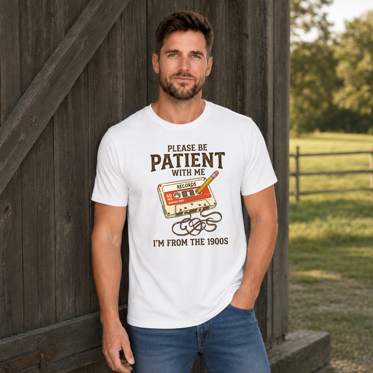 Man wearing a white T-shirt featuring the Please Be Patient With Me, I’m From the 1900s cassette design by a barn.