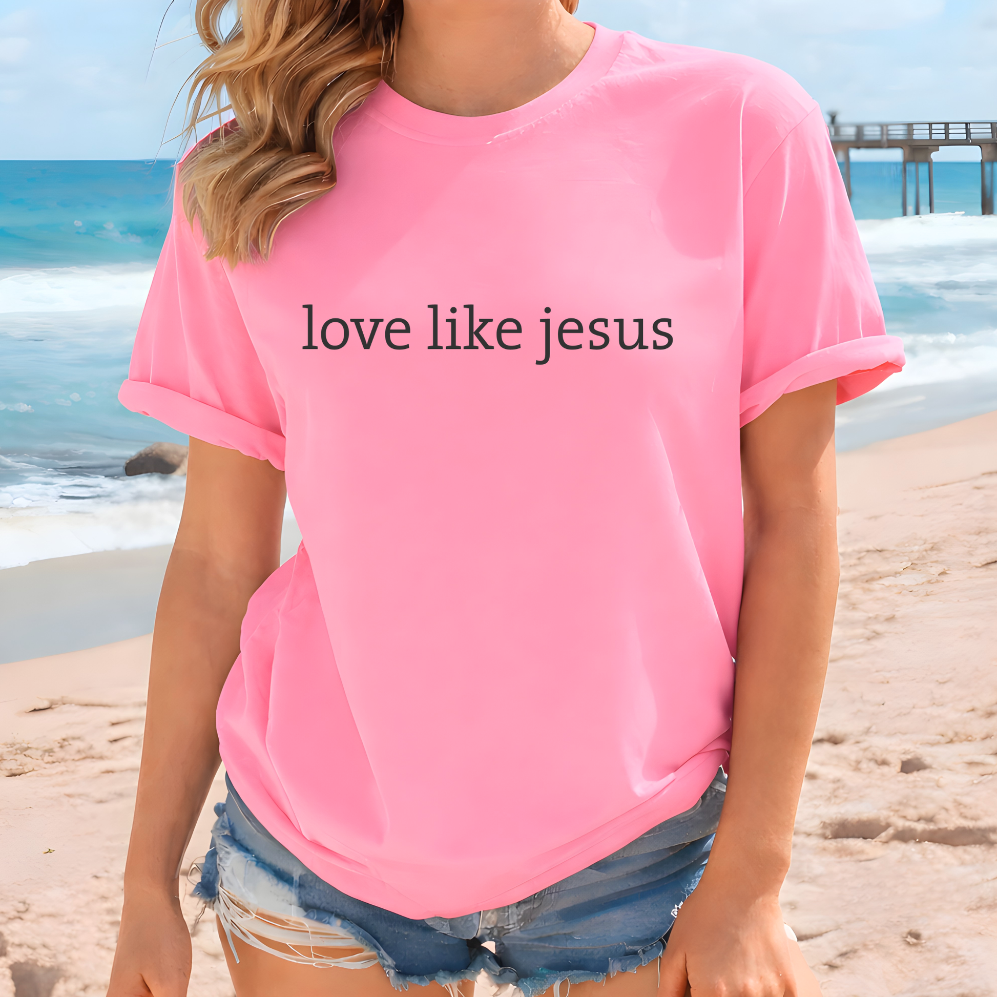 Woman wearing a pink t-shirt with 'love like jesus' text on a beach.