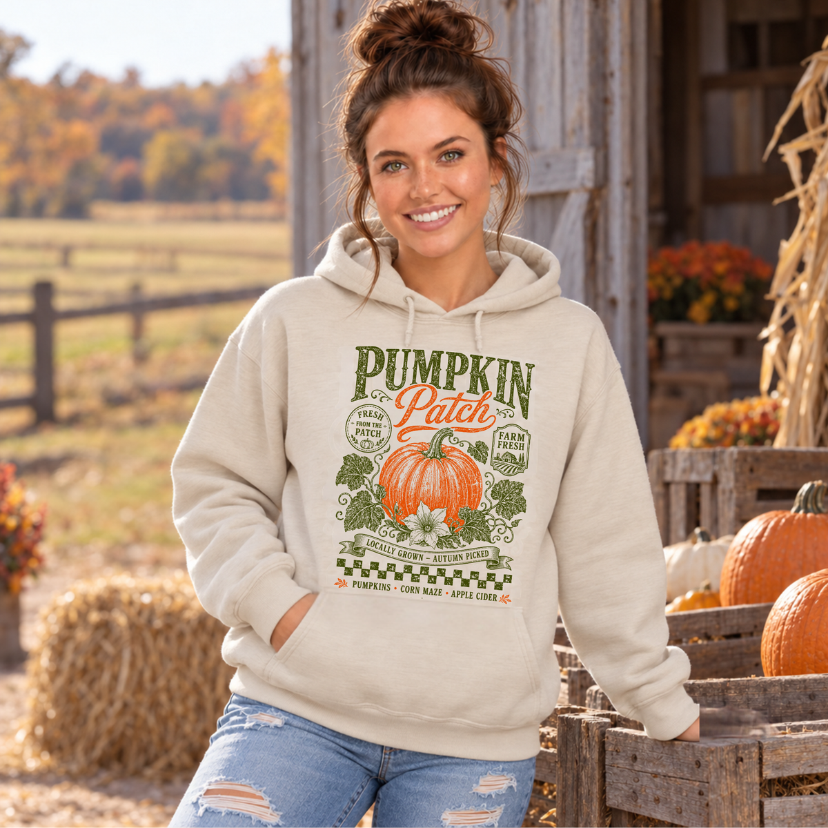 Woman wearing a sand hoodie with vintage Pumpkin Patch pumpkin and floral design beside pumpkins and hay bales
