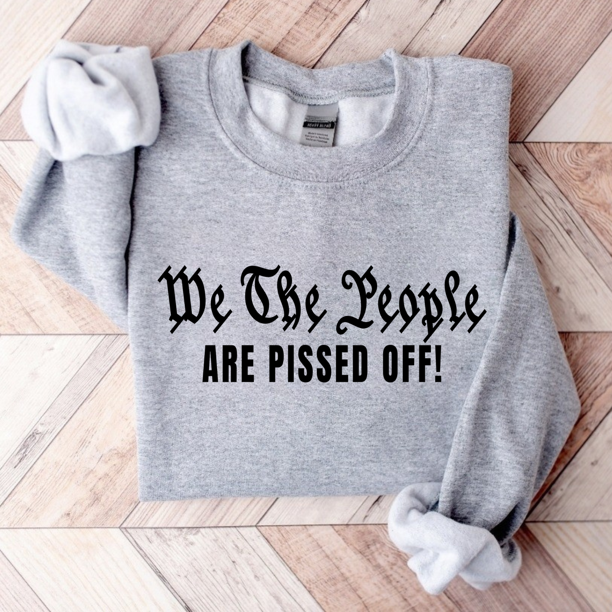 Gray sweatshirt with text 'We The People ARE PISSED OFF!' on a wooden floor.