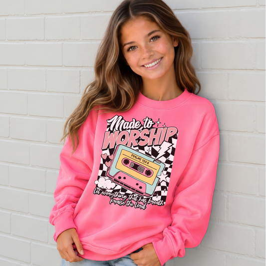 Girl wearing a Bright Pink Made to Worship youth crewneck sweatshirt with a colorful retro cassette graphic against a white brick wall.