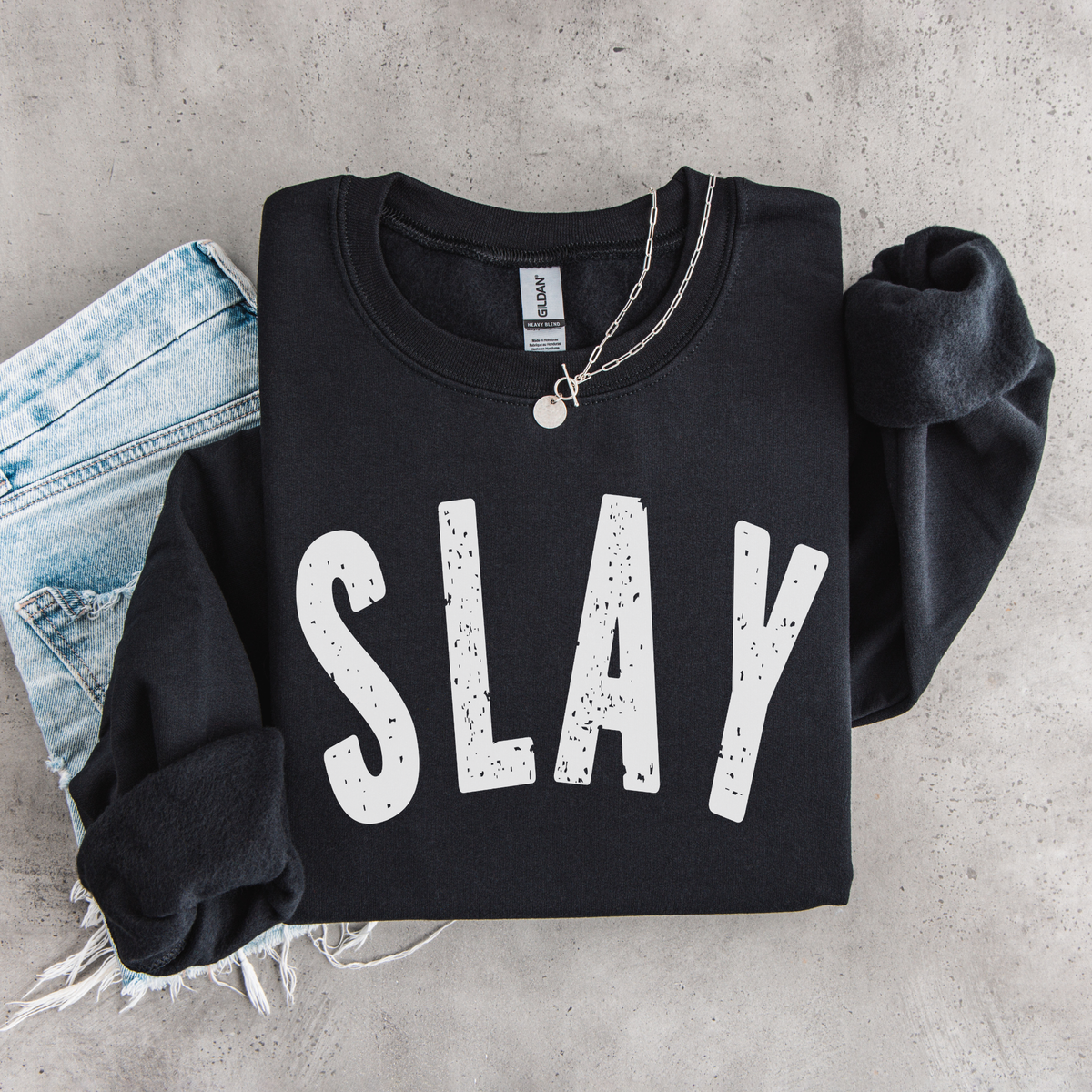 Black SLAY crewneck sweatshirt flat lay with bold white distressed lettering, styled with denim and a necklace.