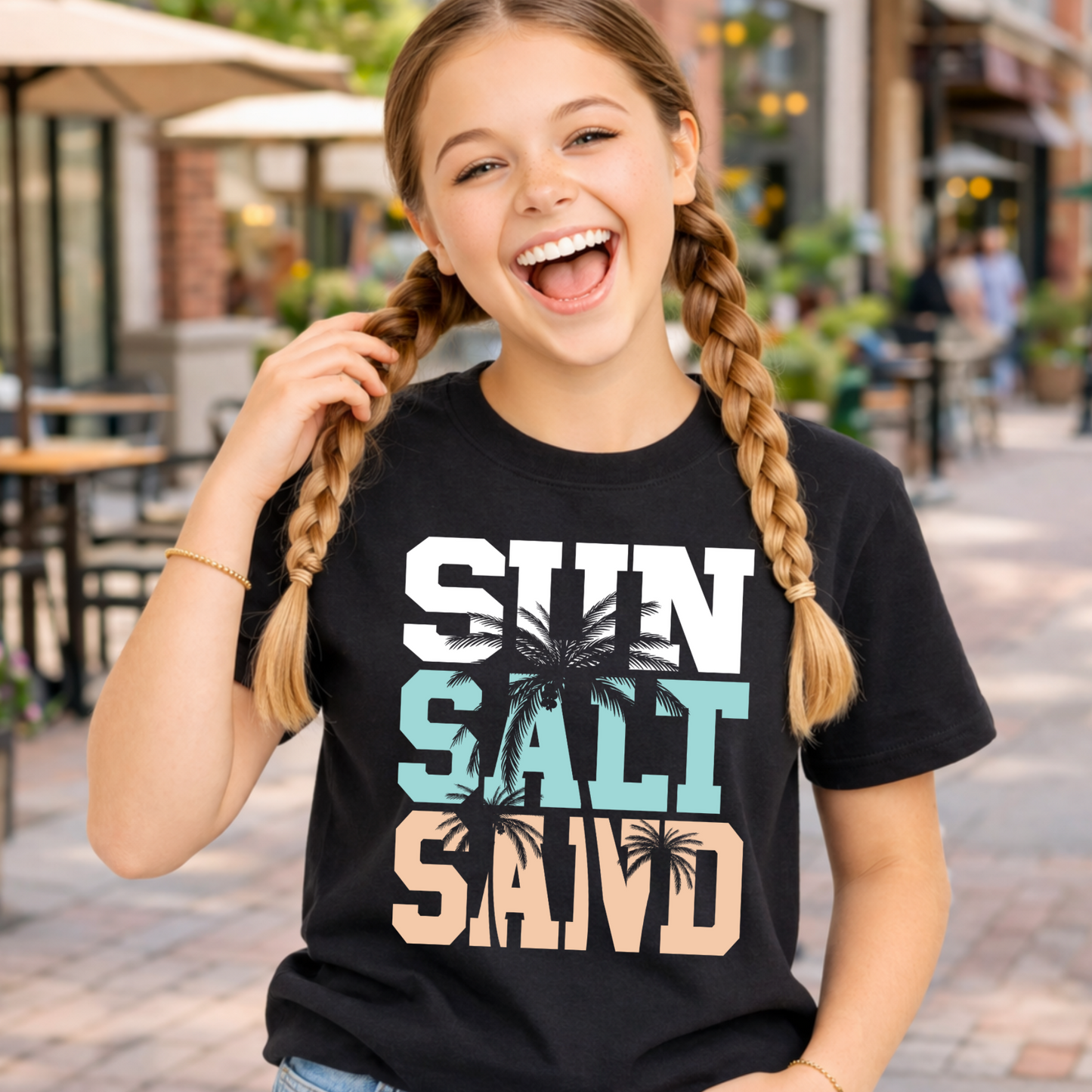 Young girl wearing a black t-shirt with 'Sun Salt Sand' text and palm tree design, standing on a street.