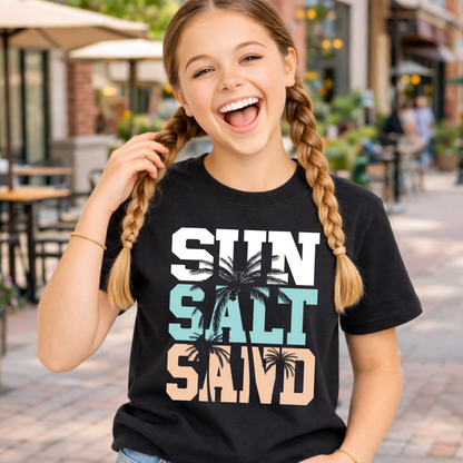 Young girl wearing a black t-shirt with 'Sun Salt Sand' text and palm tree design, standing on a street.
