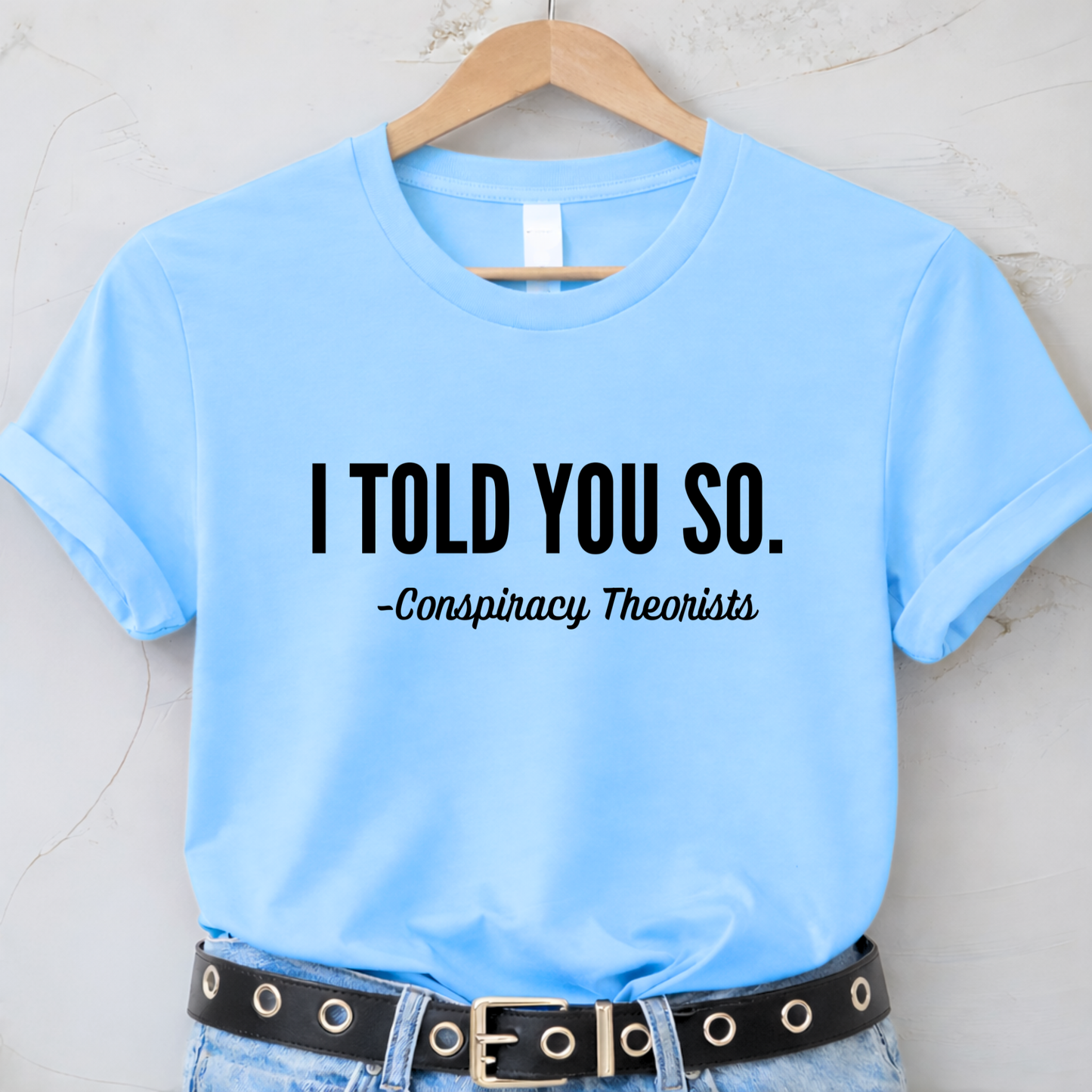 Light blue t-shirt with text 'I TOLD YOU SO. - Conspiracy Theorists' on a hanger.