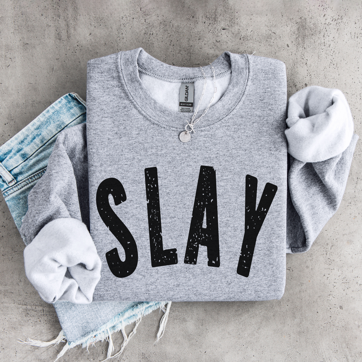 Sports gray SLAY crewneck sweatshirt flat lay with bold black distressed lettering, styled with denim, necklace, and matching scrunchies.
