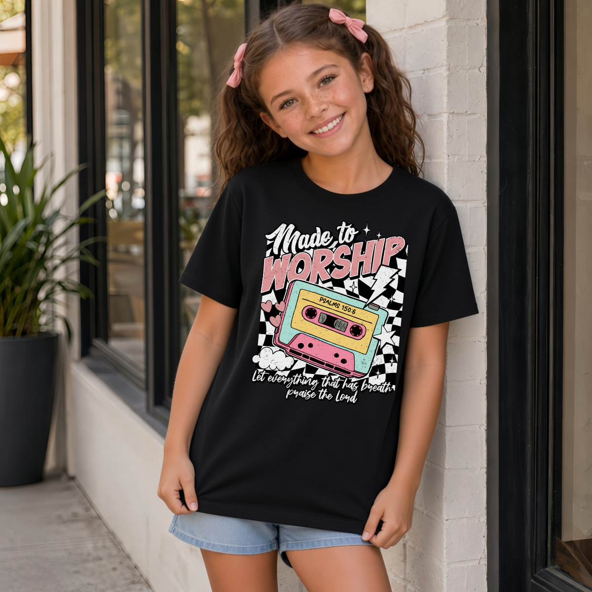 Youth girl wearing a black Made to Worship T-shirt with a colorful retro cassette worship design outside a storefront.