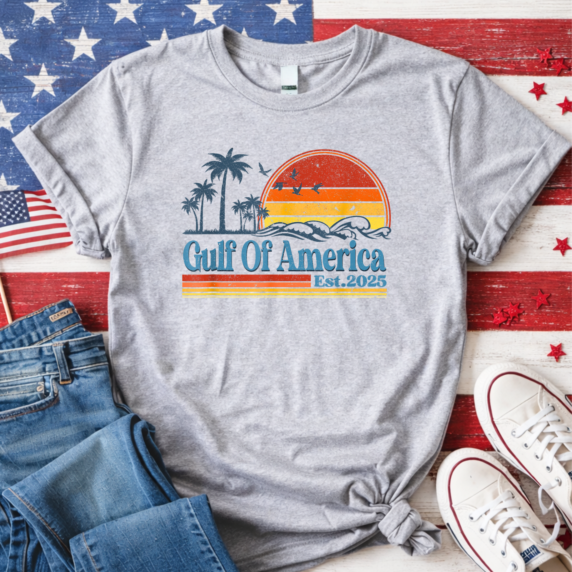 Gray t-shirt with 'Gulf of America' design on a wooden surface with American flags and shoes.