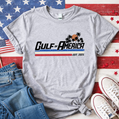 Gray t-shirt with 'Gulf of America' design on a wooden surface with American flag background