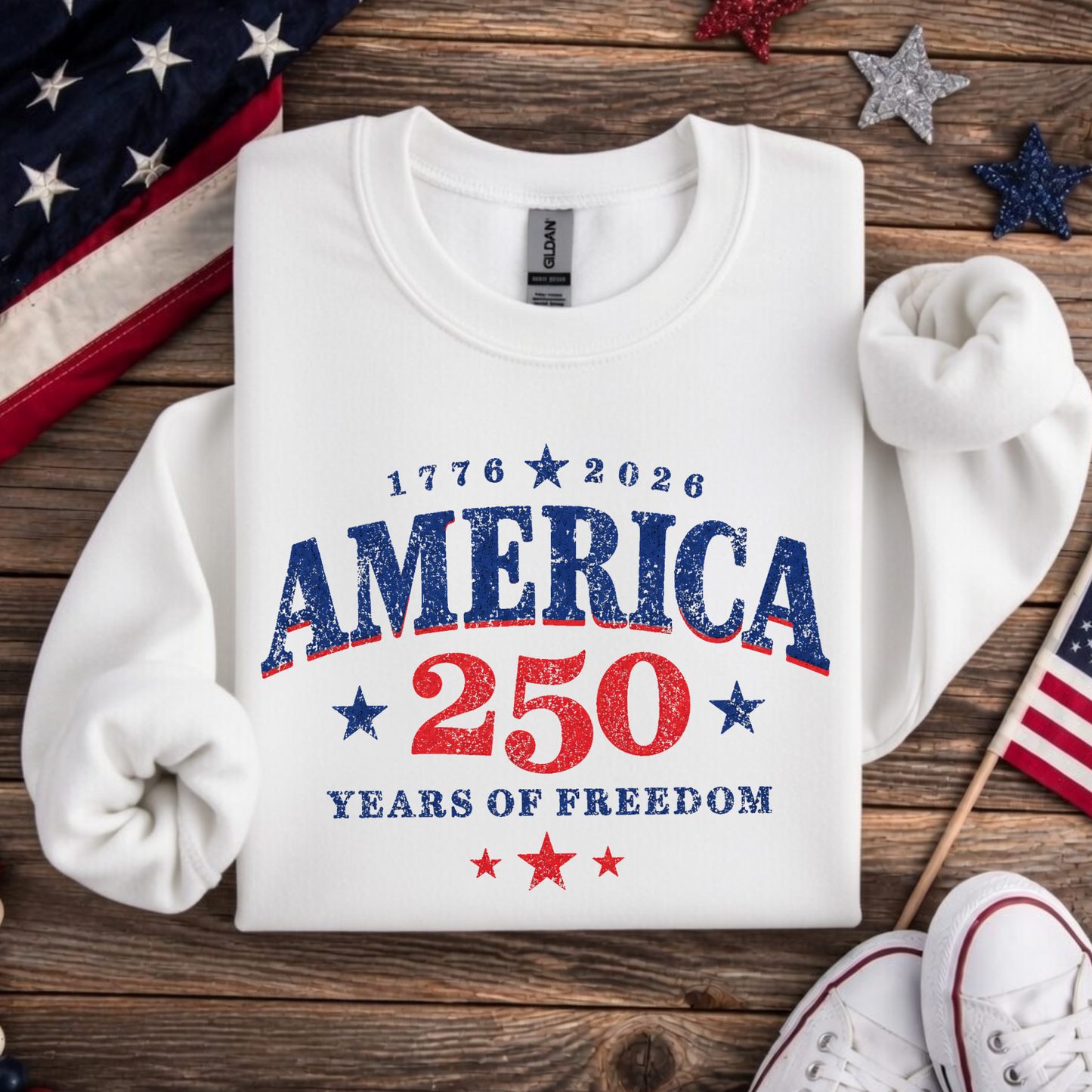 White sweatshirt with 'America 250 Years of Freedom' text on a wooden surface with American flags and stars.